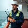 Caucasian marine deck officer chief mate on offshore vessel. Wears safety helmet, coveralls. Holds VHF radio. Stands on deck of ship. Scene shows pro seamanship safety procedures at work. Sea